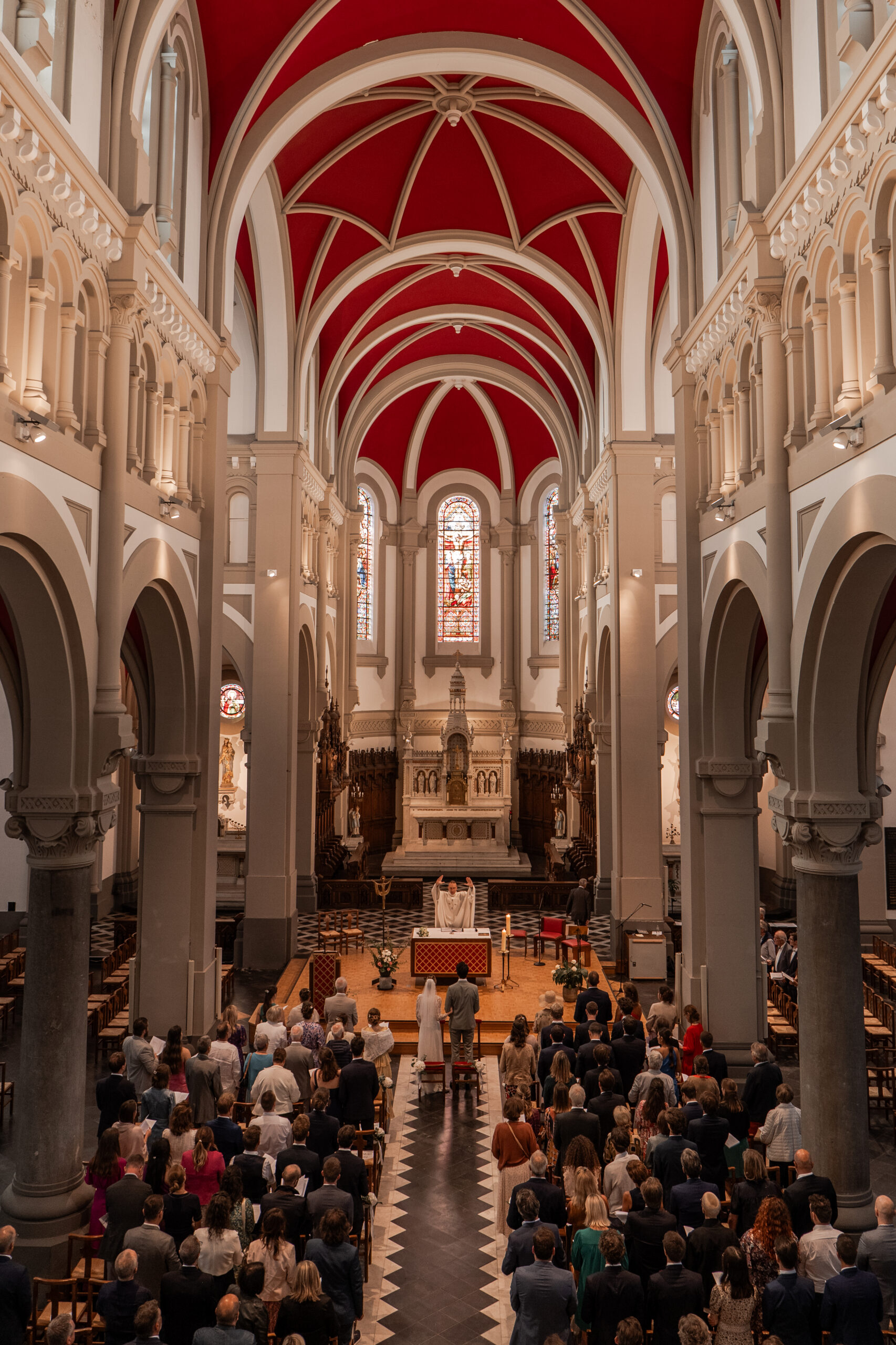 Photographie d'une cérémonie religieuse à l’église Saint Calixte à Lambersart. L'église est remplie d'invités tous debout, les mariés se trouve devant l'autel et le prêtre qui lève les bras en l'air