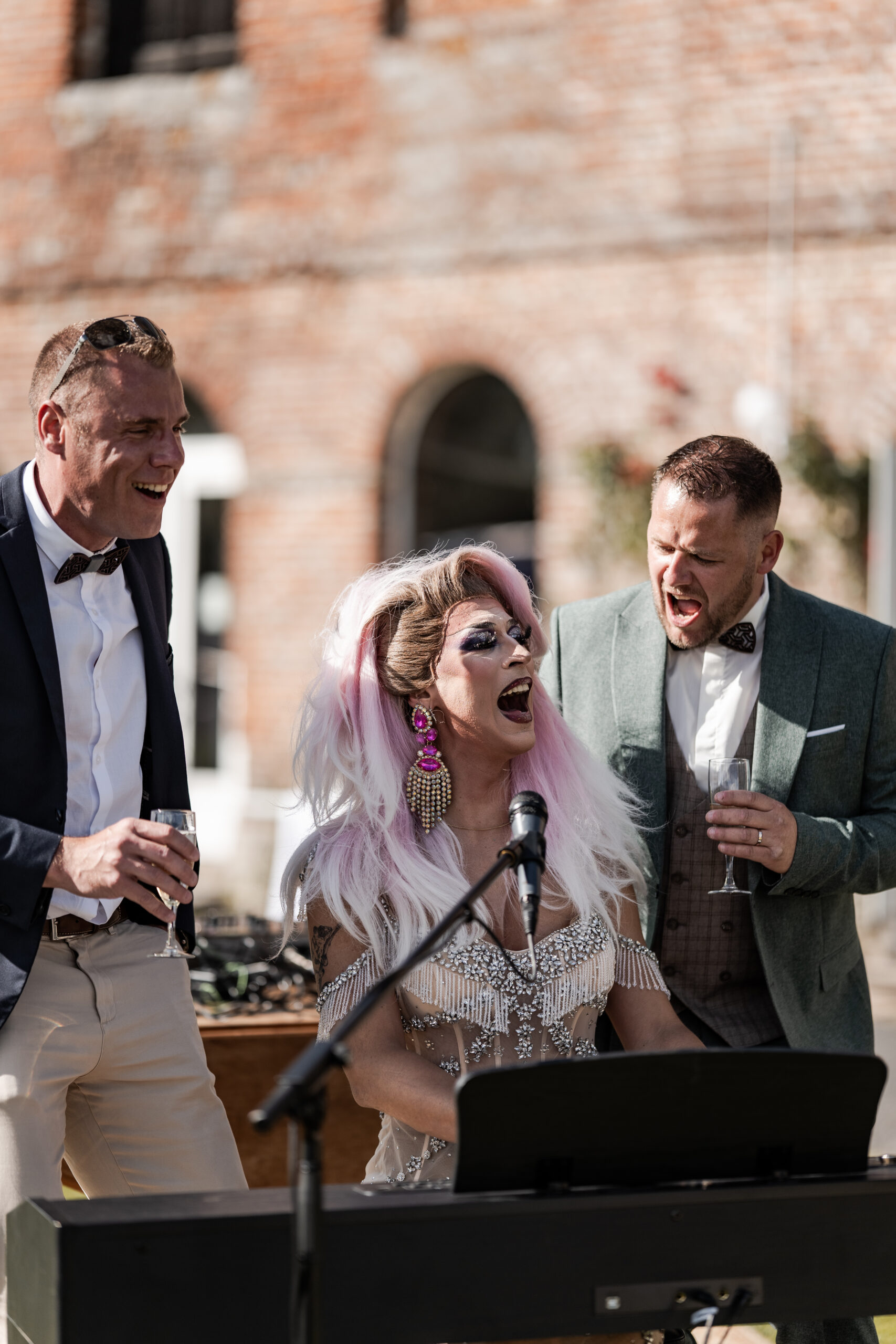 Photographie du marié avec son témoin ainsi d'une drag queen lors d'un karaoké du vin d'honneur de son mariage. Ils chantent avec un verre de champagne à la main et la drag queen joue du piano en même temps