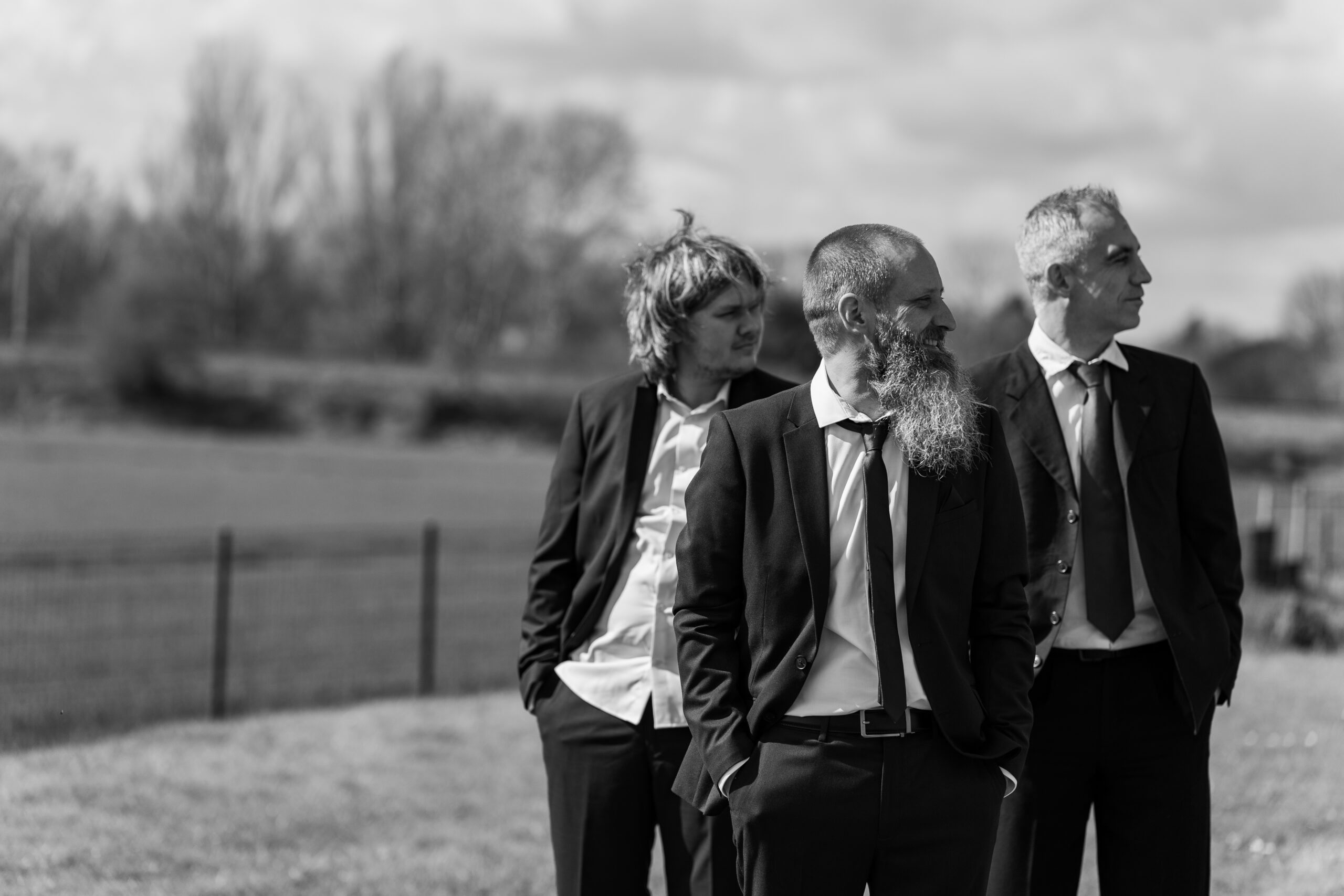 Photographie en noir et blanc d'un marié aux côtés de ses deux témoins les mains dans les poches et regardant vers le côté lors des préparatifs de son mariage