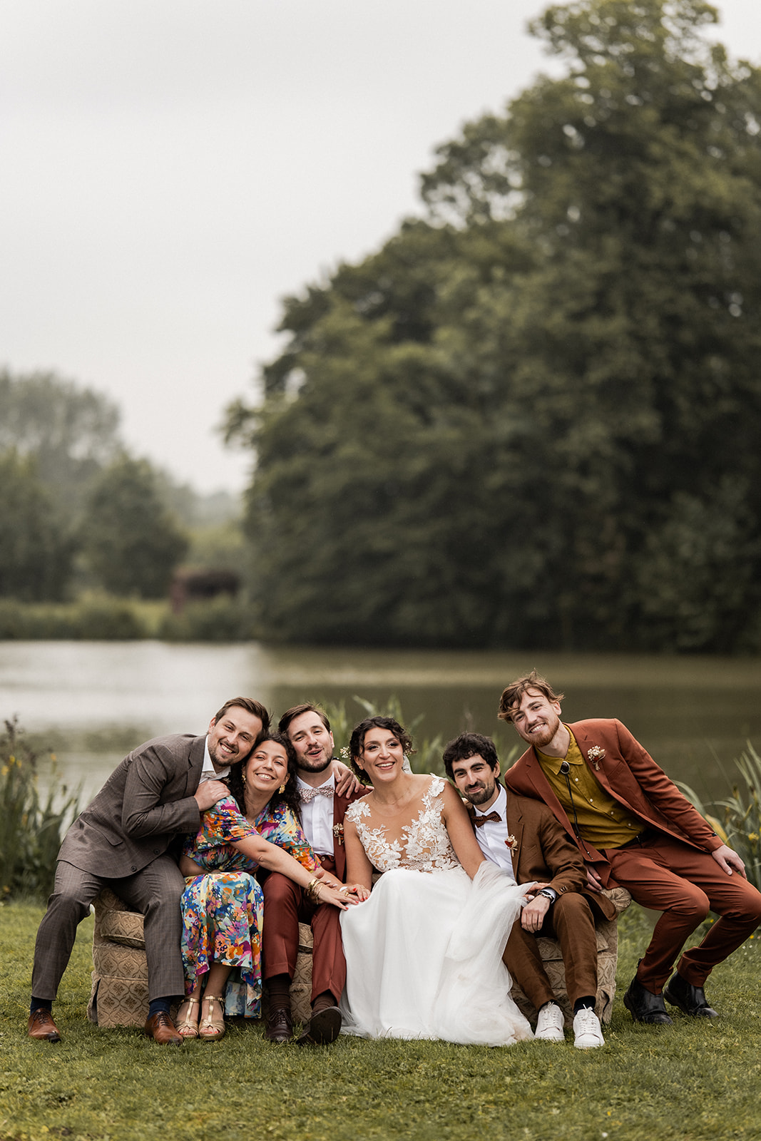 Photographie d'un couple de mariés avec quelques amis lors d'une séance photo de goupes dans le jardin de la Cense d’Abaumont en Belgique devant un étang. Ils sont assis sur un canapé vintage et sourient en regardant la caméra