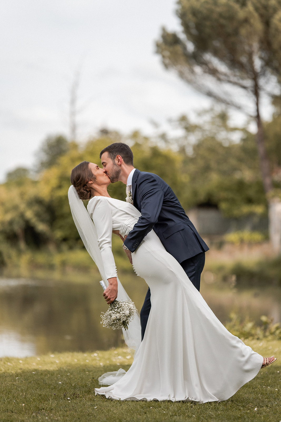 Photographie d'un couple de mariés lors de leur séance photo de couple de leur mariage. Le marié renverse la mariée en arrière pour l'embrasser, devant un petit étang à Villers-Cotterêts
