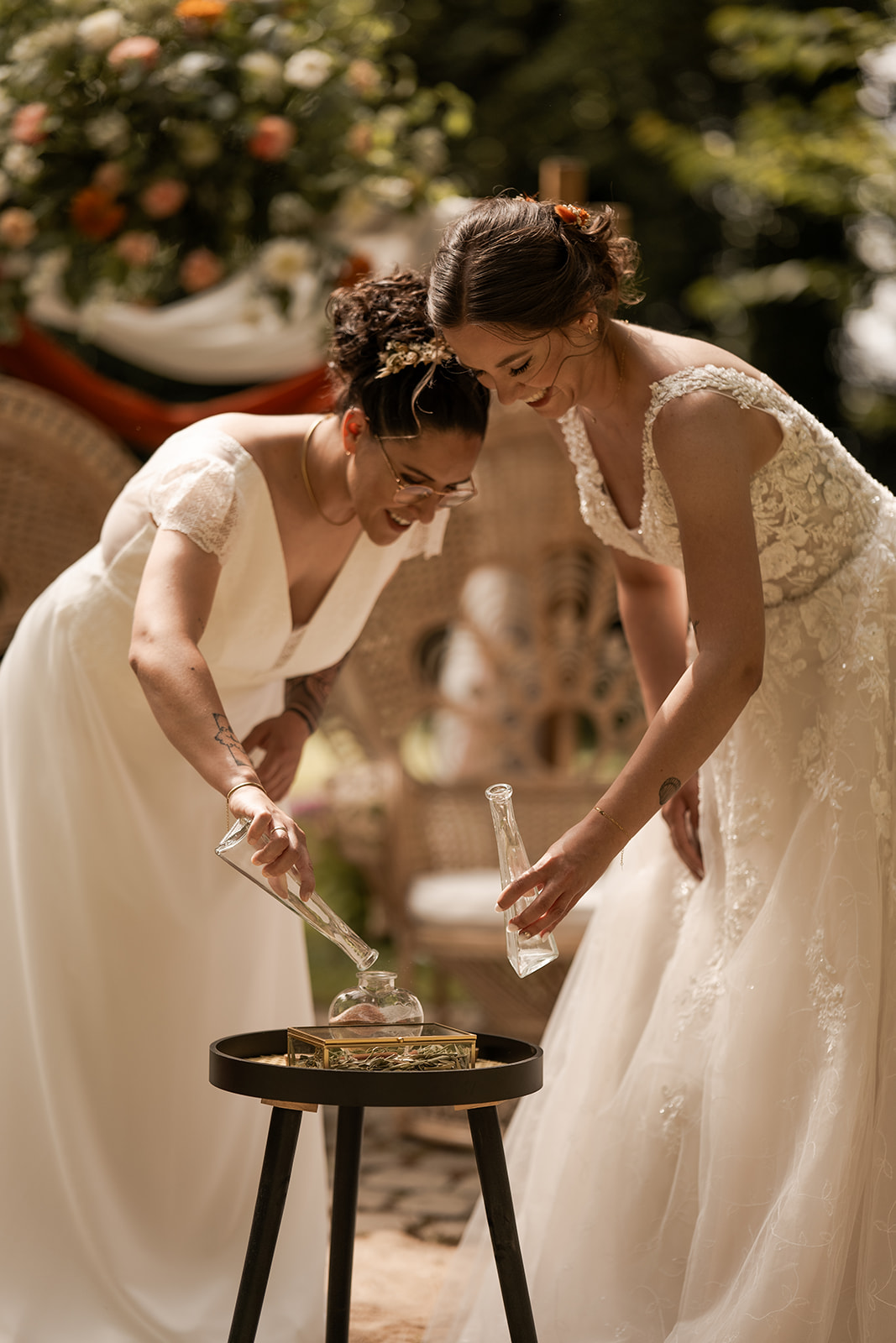 Photographie de deux mariées pendant leur cérémonie laïque faisait leur rituel du sable. Elles portent toutes les deux une robe blanche et se baissent pour remplur un petit bocal de sable