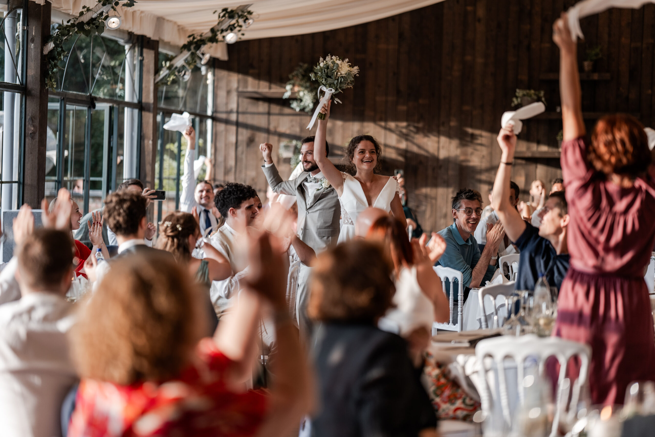 Photograhie de l'entrée en salle d'un couple de mariés à l’orangerie de Beauval en Belgique entourés de leurs invités qui lèvent les bras en l'air et font tourner des serviettes de tables en l'air en les acclamant