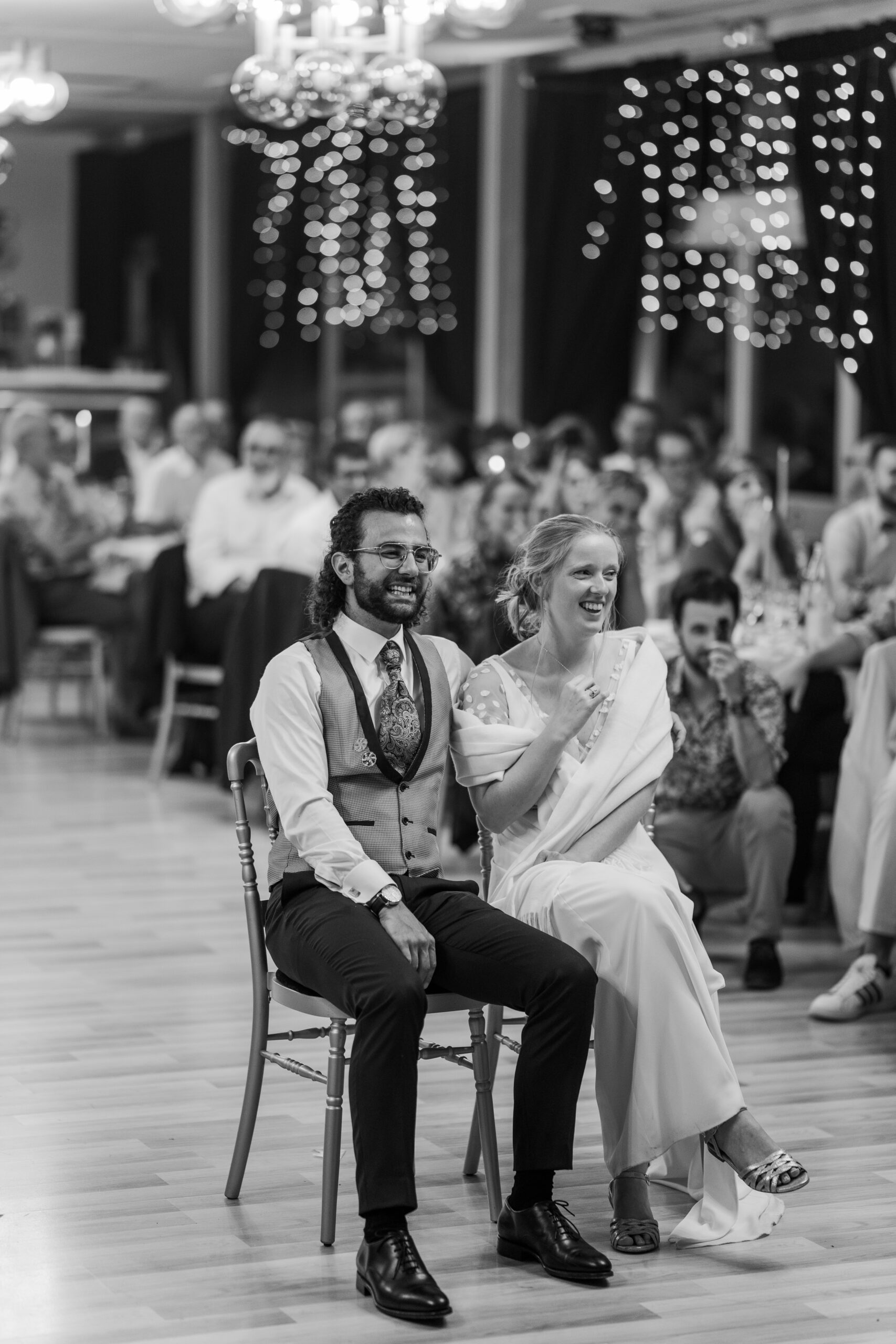 Photographie en noir et blanc d'un couple de mariés assis et qui rigole en regardant leur témoins hors photo qui font un discours pour leur mariage