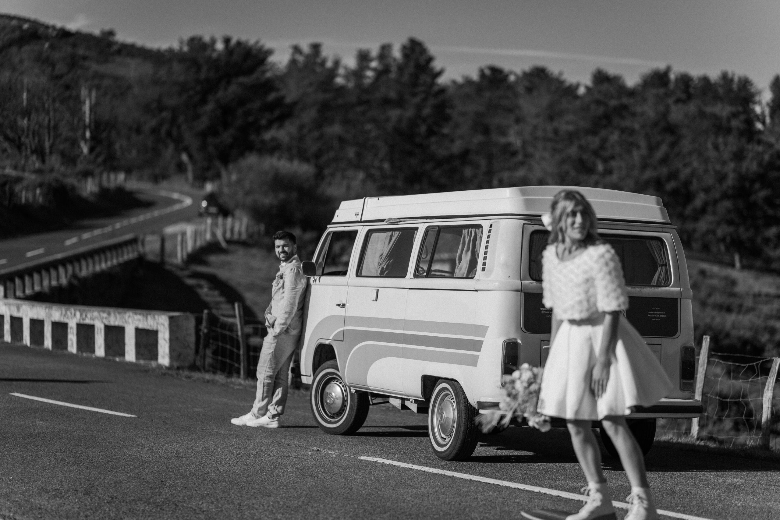 Photographie en noir et blanc d'un couple de mariés sur la route d'une montagne dans les Pyrénées aux côtés d'un van vintage, la marié en robe blanche fait du skateboard et le marié la regarde adossé contre le van