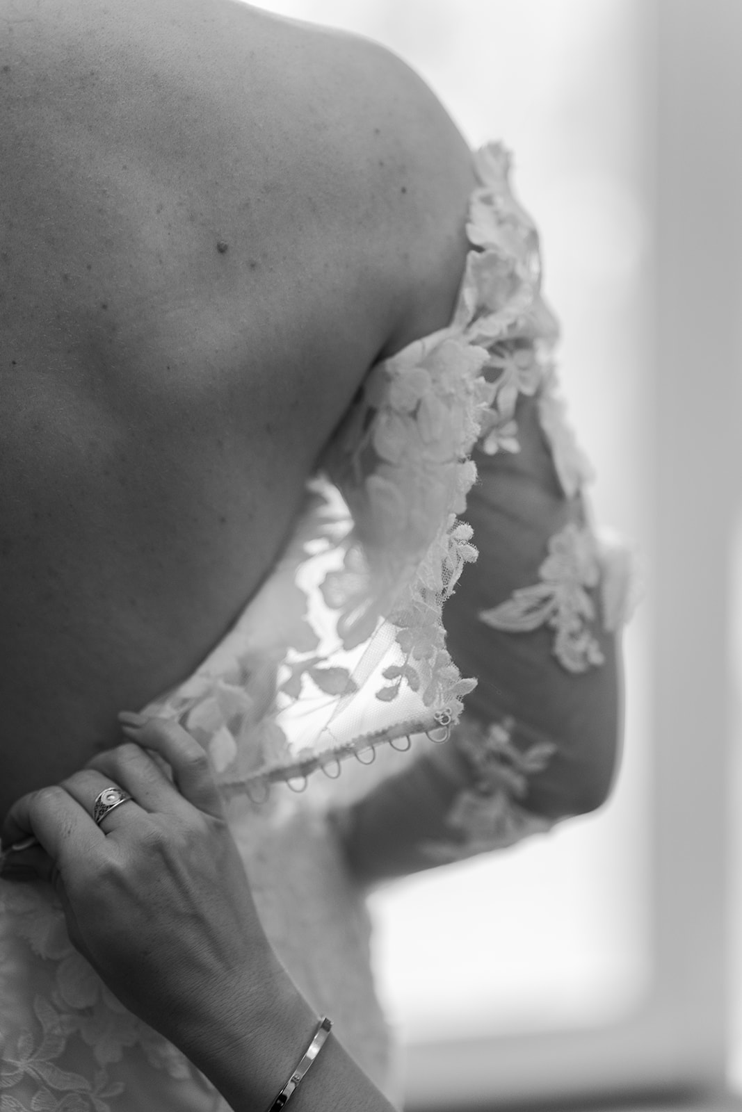 Photographie en noir et blanc du dos d'une mariée qui se fait fermer sa robe de mariée par une femme dont on ne voit qu'une main