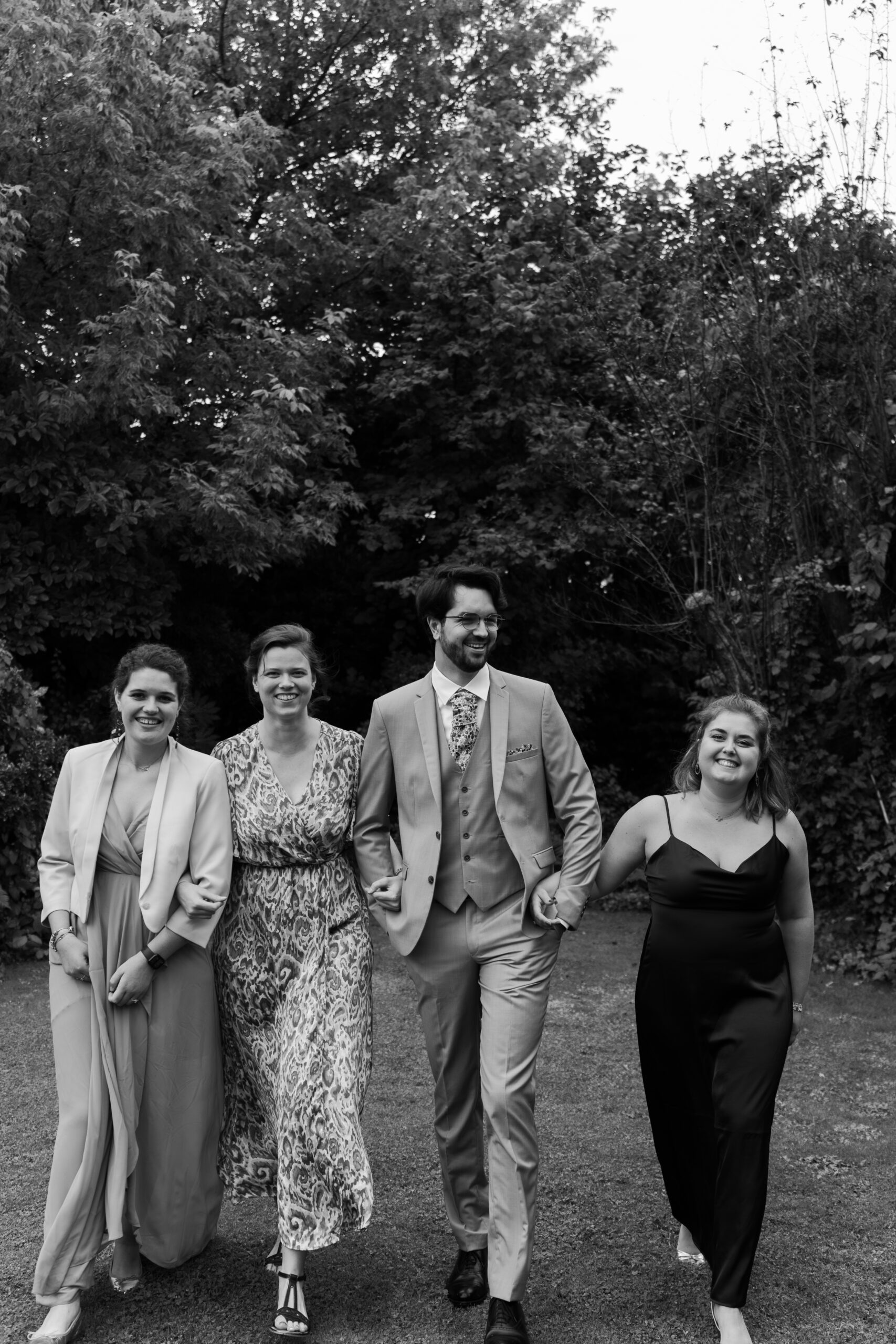 Photographie en noir et blanc du marié bras dessus bras dessous avec 3 amies marchant vers l'appareil photo et rigolant