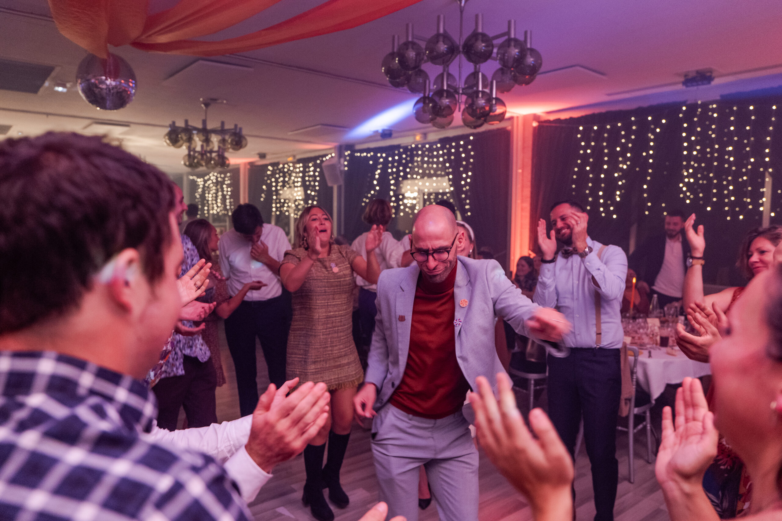 Photographie d'un homme qui danse au milieu d'un cercle d'invités pendant la soirée dansante d'un mariage à thème années 70