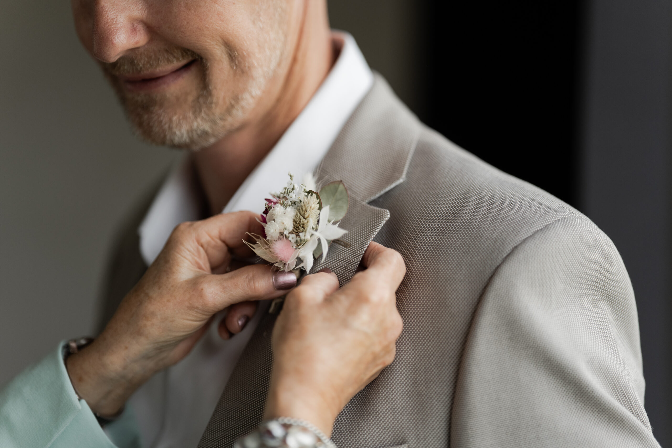 Photographie d'un homme qui se fait poser sa broche en fleurs sur sa veste par sa mère dont on ne voit que les mains lors des préparatifs de son mariage