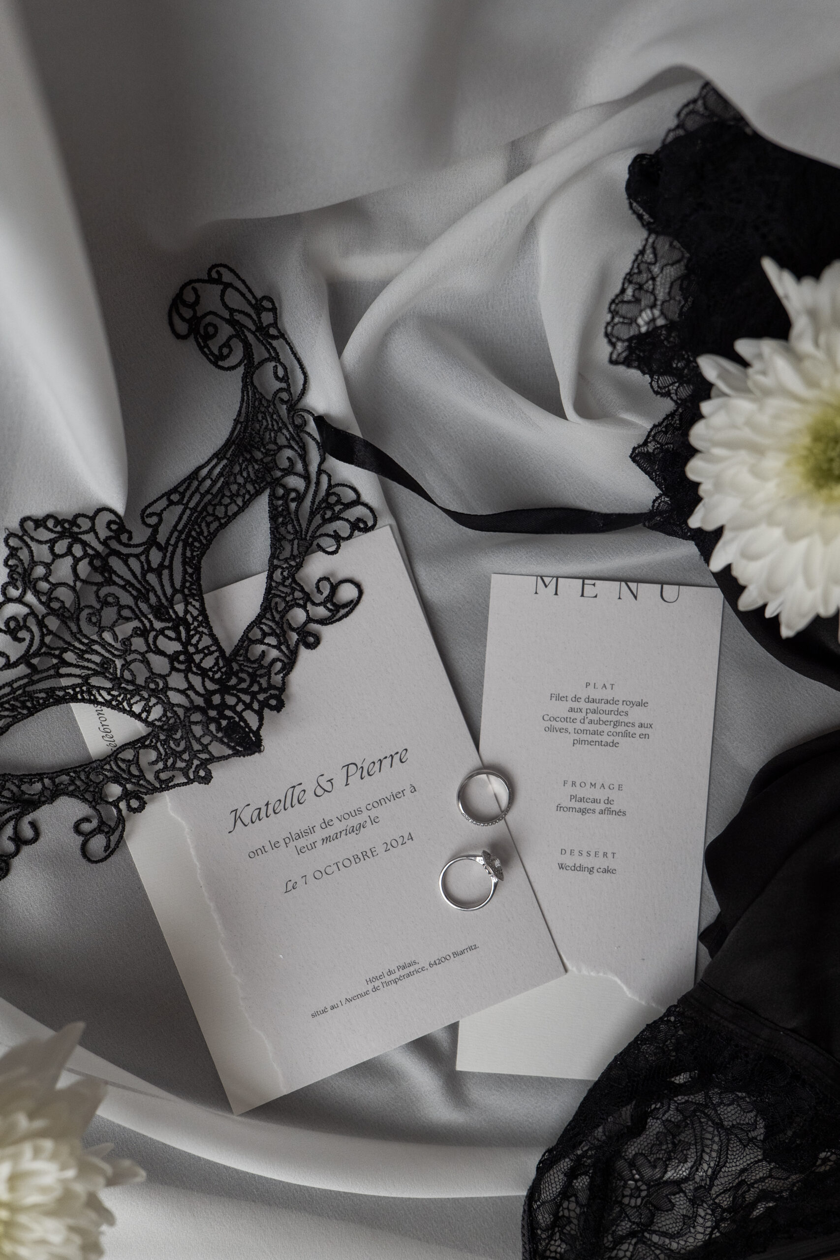 Photographie d'un flatlay de faire part avec les alliances quelques fleurs et un masque en dentelle