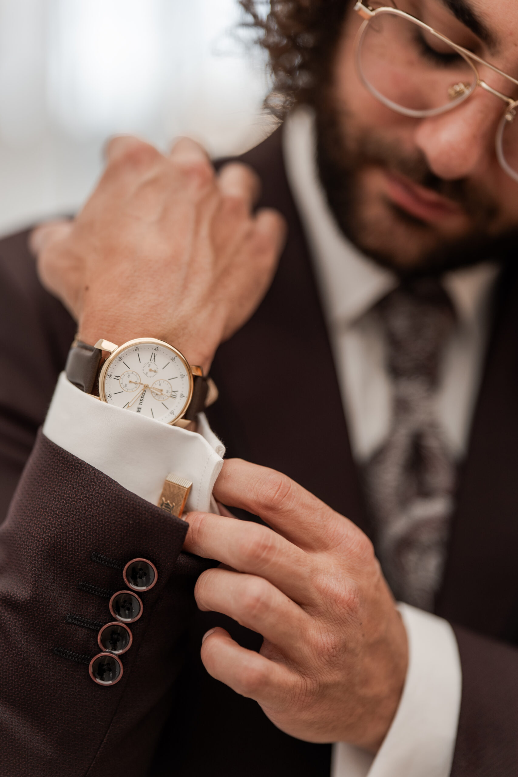 Photographie de détails du mariés lors des préparatifs de son mariage: l'homme est flou mais le focus est sur ses mains et sa montre qu'il ajuste à son poigné
