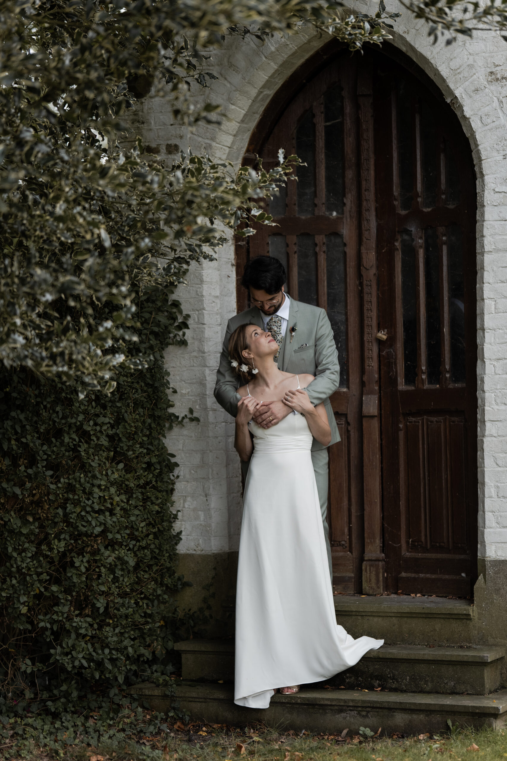 Photographie d'un couple de mariés pendant leur séance couple de leur mariage, il est en costume et elle est en robe blanche, le marié l'enlace et ils se regardent