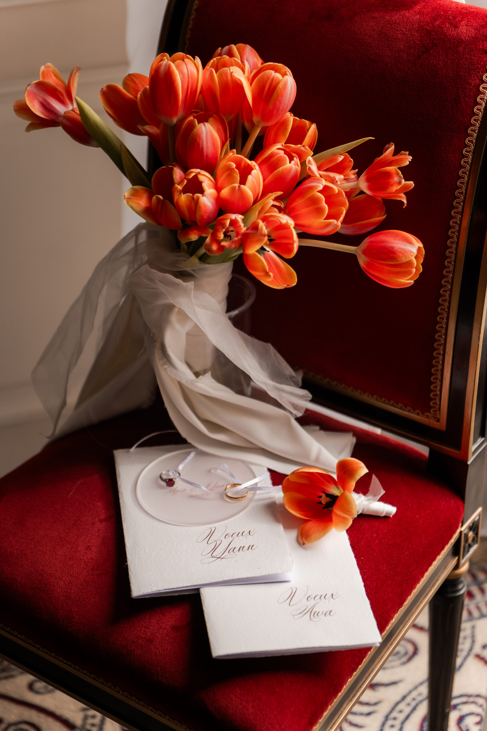Photographie d'un flatlay des faire part de mariage ainsi que les alliances et un bouquet de tulipes orange posés sur une chaise en velours rouge