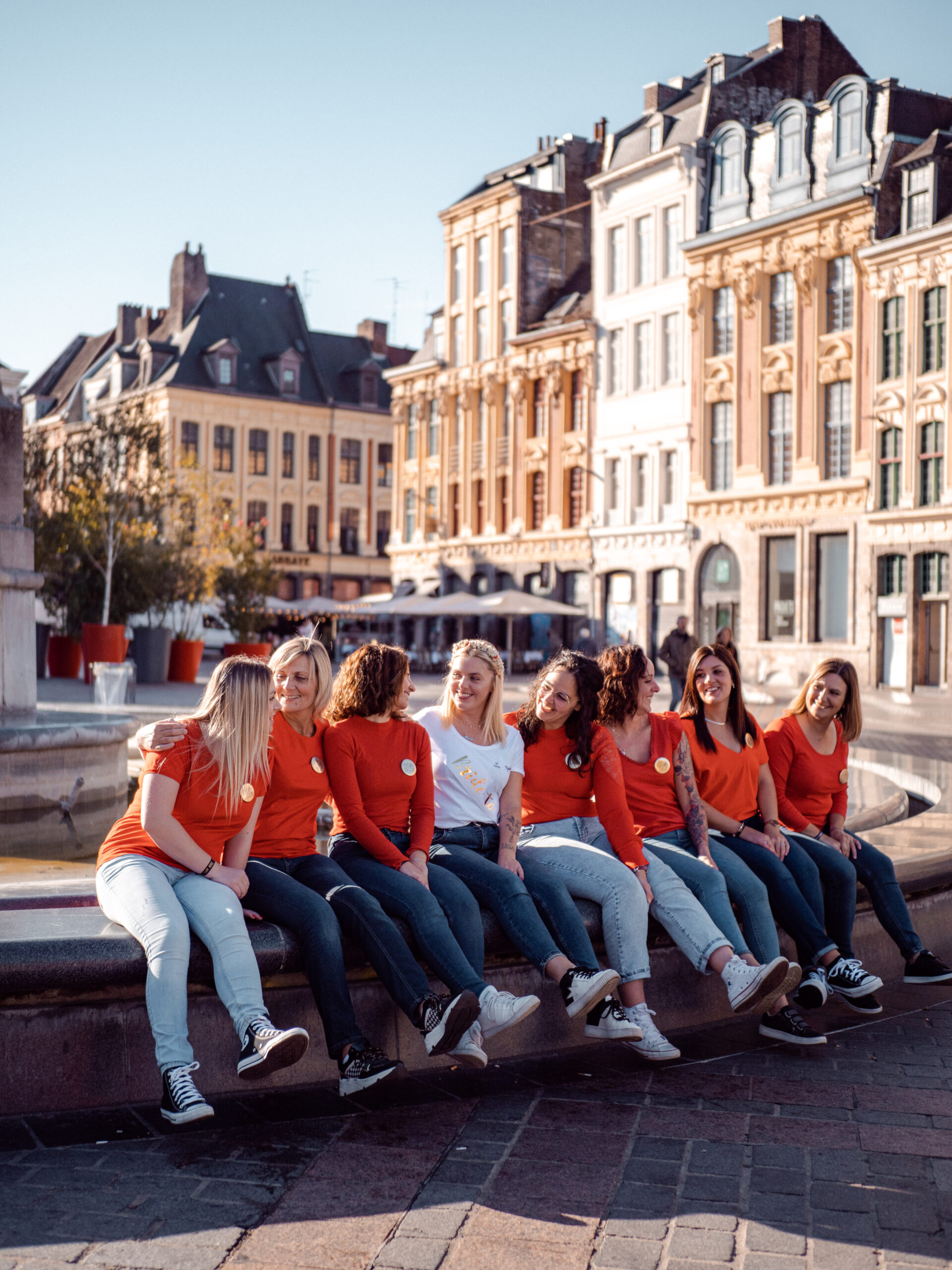 Photographie d'un groupe d'amies assises sur la fontaine de la Grande Place de Lille toutes avec un haut rouge sauf la future mariée au milieu lors d'un EVJF