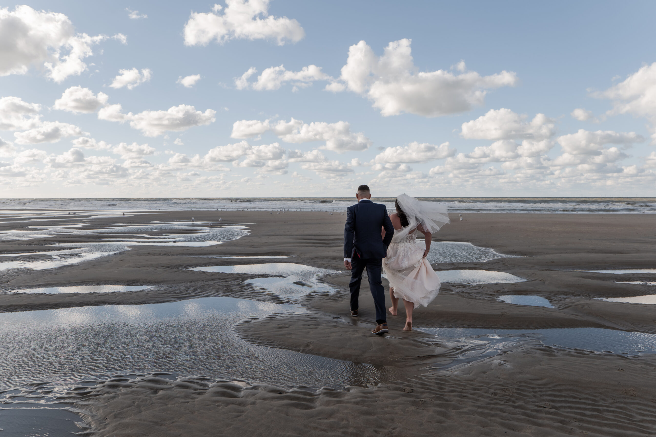 Photographie d'un couple de mariés tournant le dos au photographe et marchant pieds nus sur le sable de la plage d'Ostende pendant la marée basse