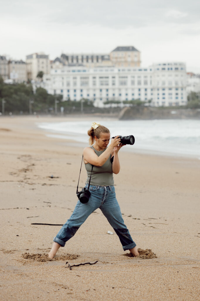 Photographie de la photographe du site internet qui a les pieds dans le sable sur la plage de Biarritz, prenant une photo au loin aillent un air très concentré