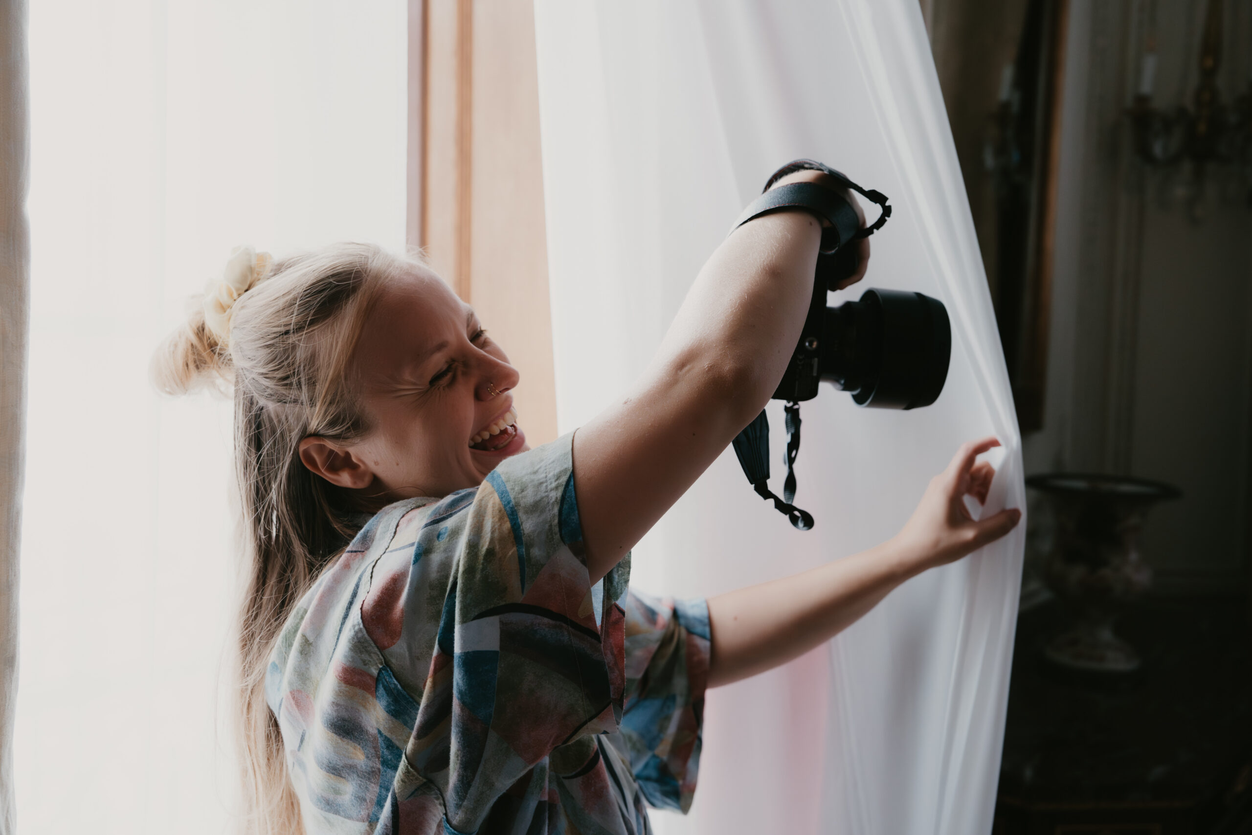 Photographie de la photographe du site internet rigolant et prenant une photo dans l'encadrement d'une porte repoussant doucement le drapé de la porte