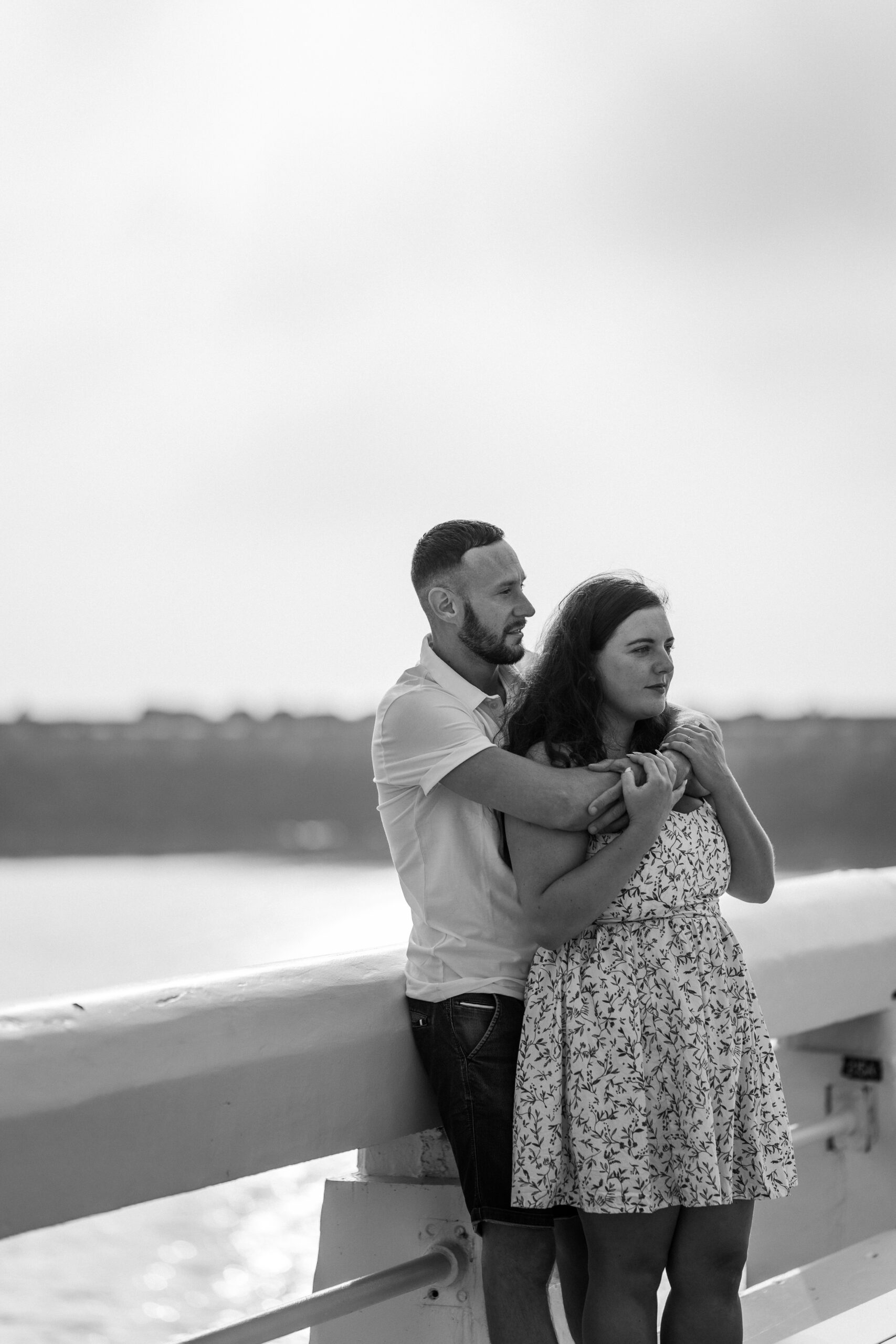 Photographie en noir et blanc d'un couple de futurs mariés enlacés dans les bras l'un de l'autre sur le ponton de la plage d'Ostende lors d'une séance photo couple