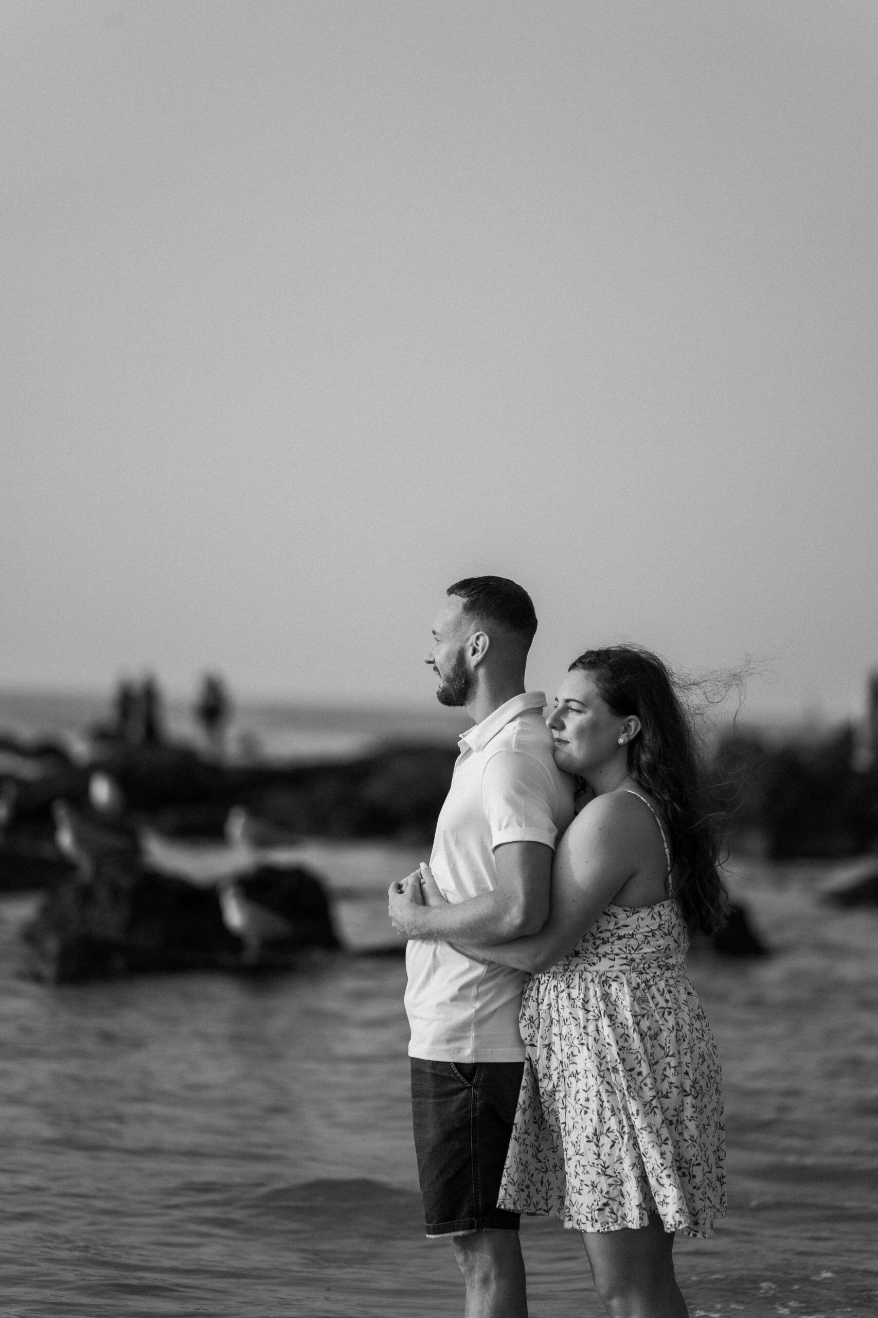 Photographie en noir et blanc d'un couple de futurs mariés enlacés dans les bras l'un de l'autre les pieds dans l'eau sur la plage d'Ostende lors d'une séance photo de couple