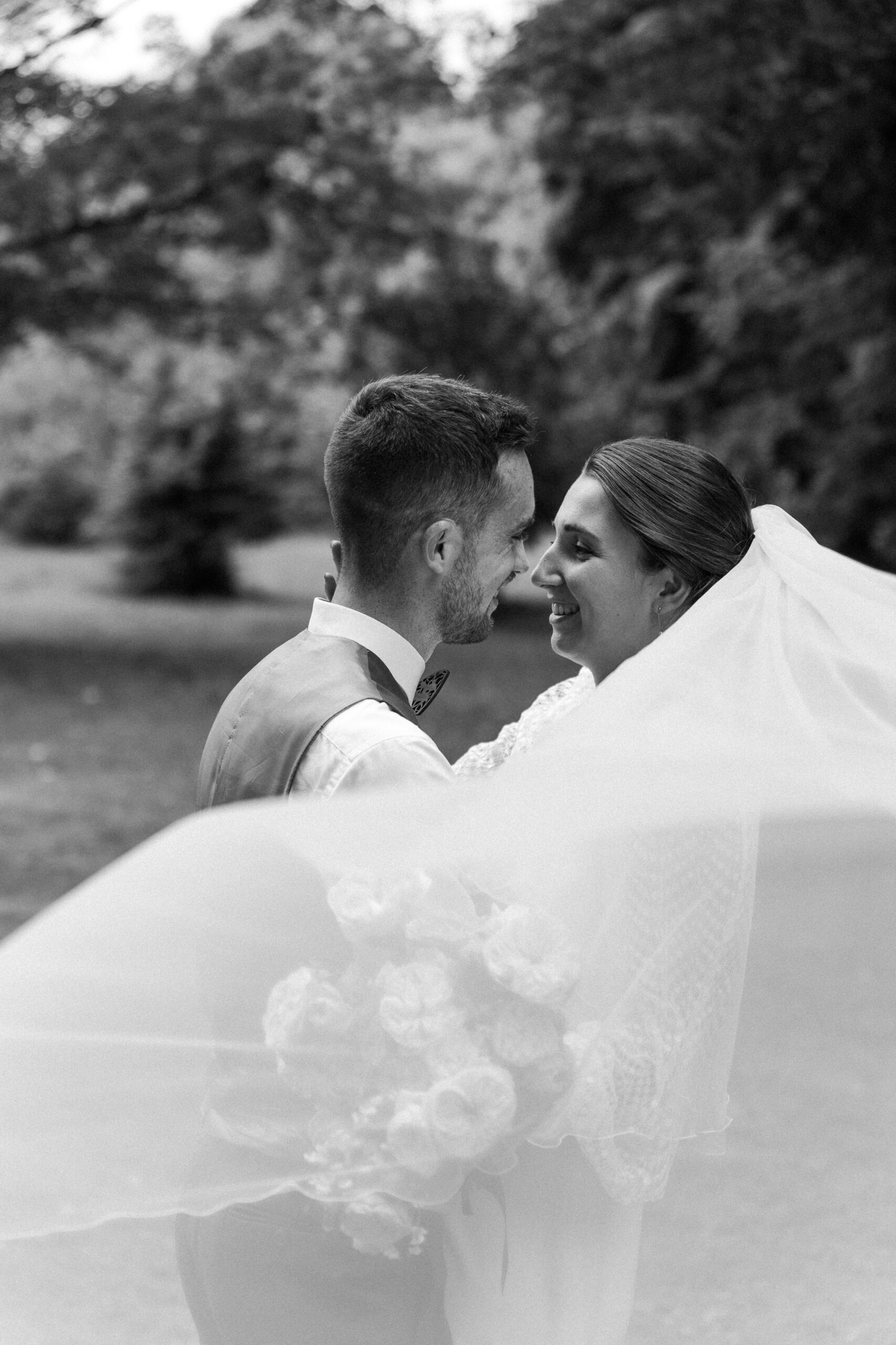 Photographie en noir et blanc d'un couple lors de leur mariage se regardant le voile volant devant l'objectif